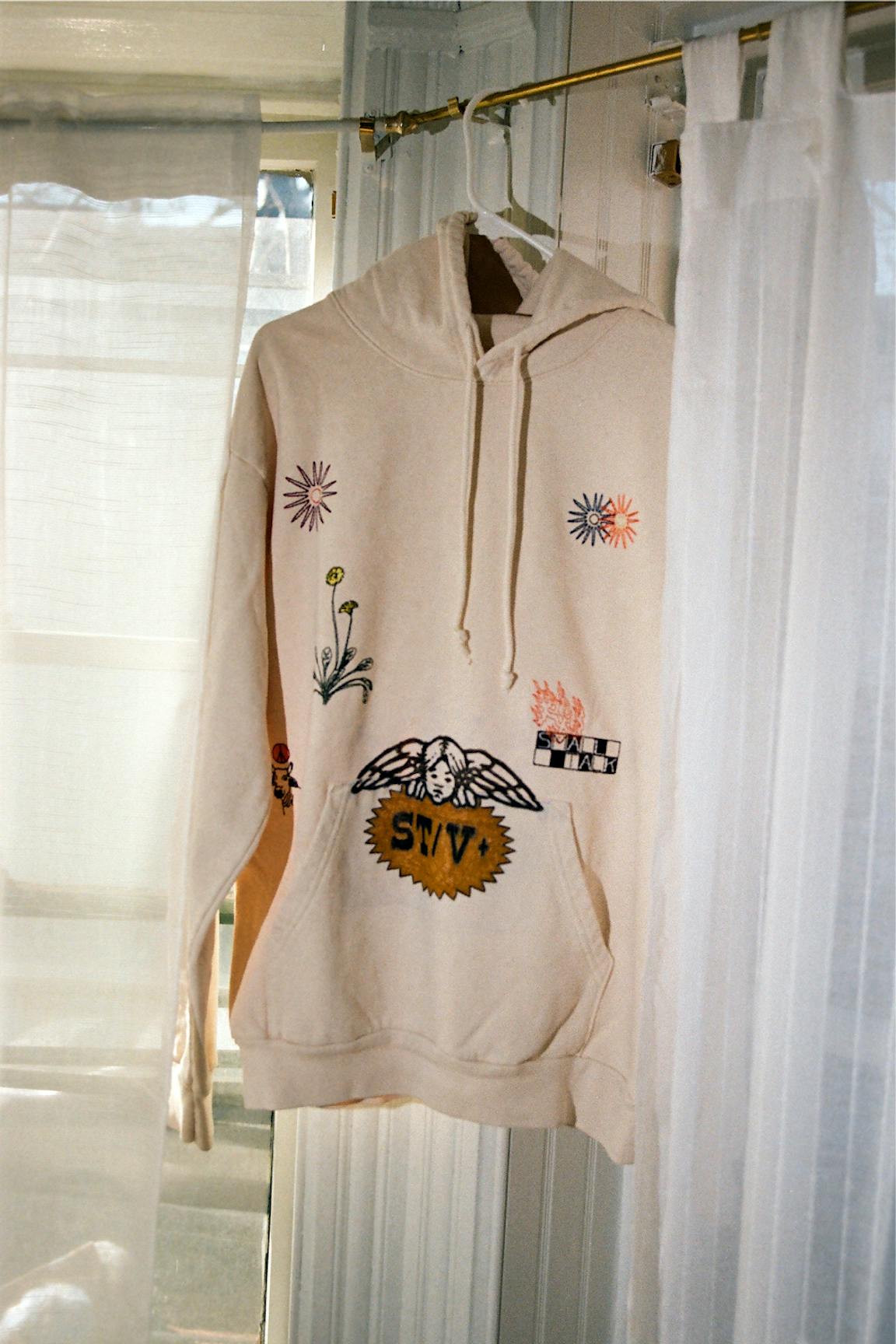 An illustrated and embroidered hoodie hangs in front of a window.